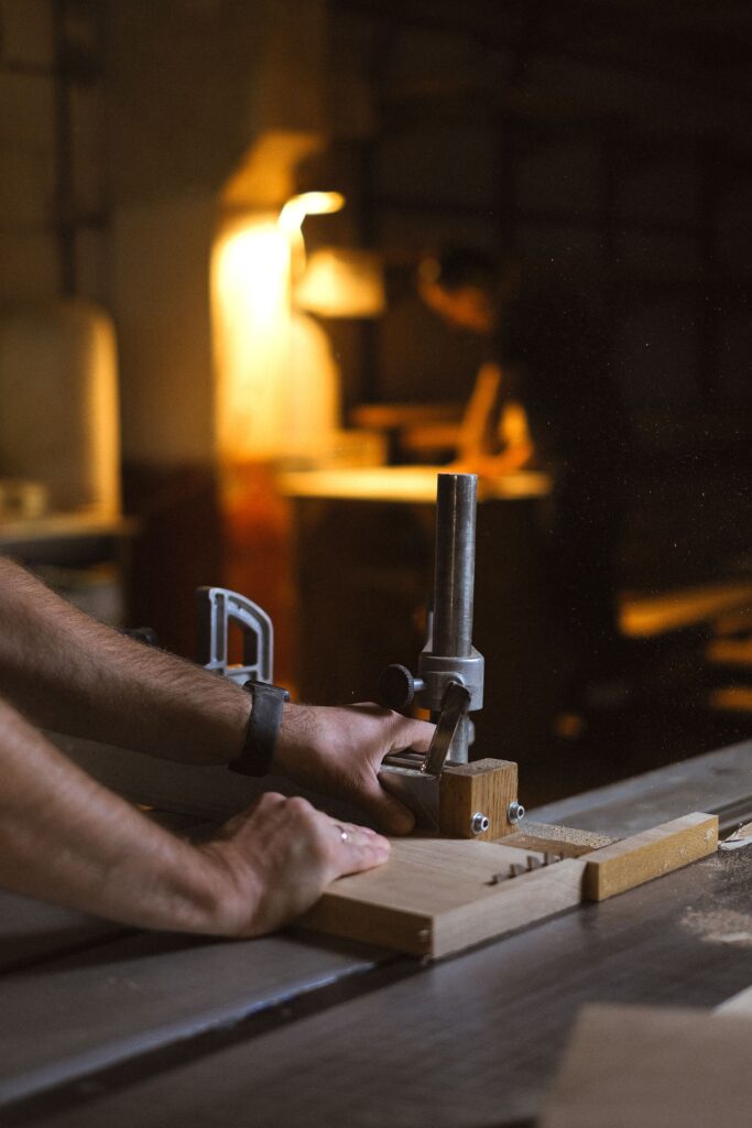 Unrecognizable male master using circular saw to cut wooden plank while working in professional joinery with glowing lamp on blurred background